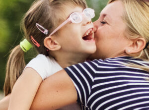 A vision impaired preschool girl is with her Mum in the park. She is laughing as her Mum hugs and gives her a kiss.