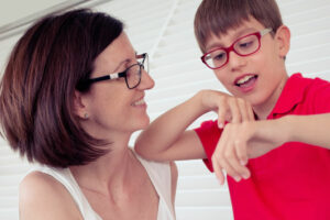 Mother and son are standing in front of venetian blinds. She is looking at him lovingly as he plays with his hands.