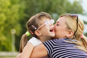 A vision impaired preschool girl is with her Mum in the park. She is laughing as her Mum hugs and gives her a kiss.