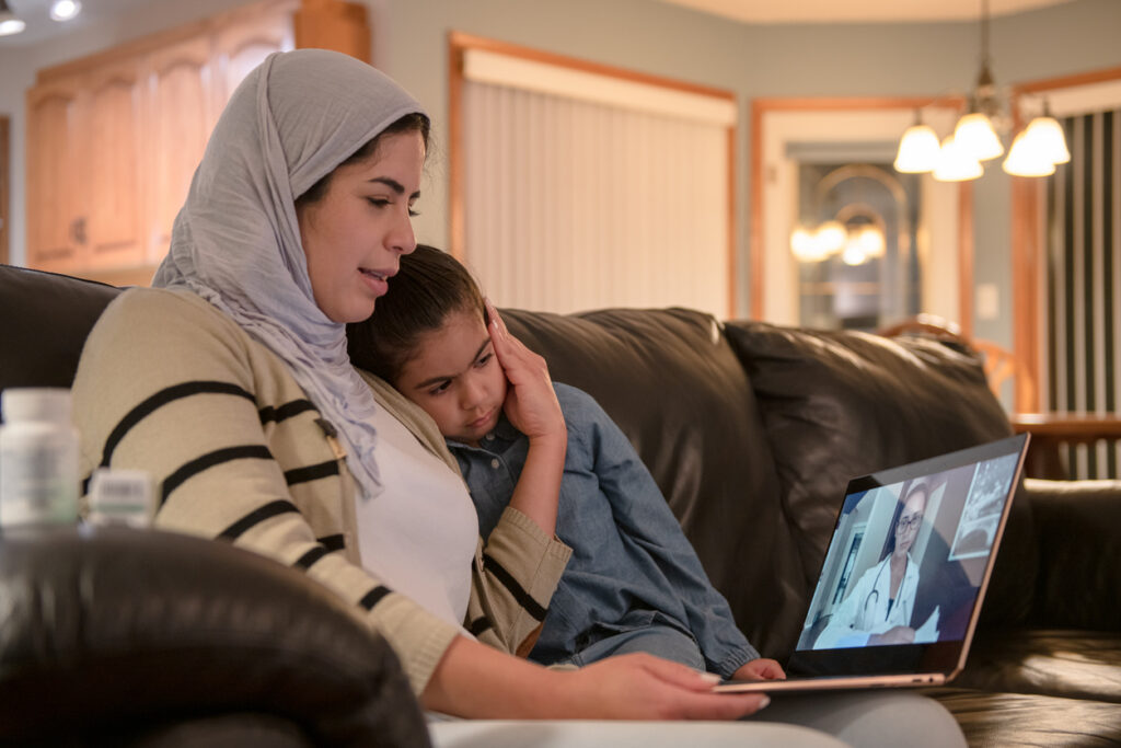 A woman and her daughter speak to a doctor via video call on a laptop. The mother has a hand on her daughters face.