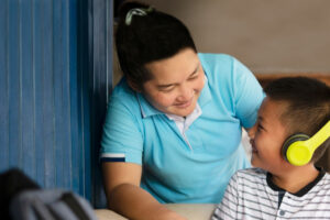 A young boy wearing headphones sits on a couch. He is using a laptop and is looking up at his mother. They are smiling at each other.