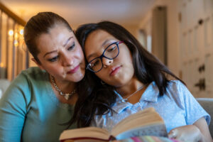 Teenage girl is with her Mum at home. The daughter wears glasses and leans her head into her Mum. They read a book.