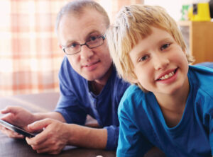 A father lays on the floor with his teenage son. He wears glasses, and they smile. The father holds an iPad.
