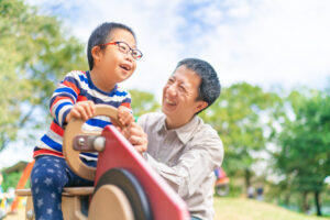 A young Asian boy with Down syndrome plays with his Dad in the park. They have fun as the boy pretends to drive a car.