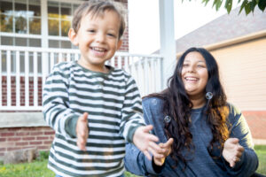 A young boy with disability is with his mother. She is smiling and blowing bubbles. He is holding out his hands to catch them.