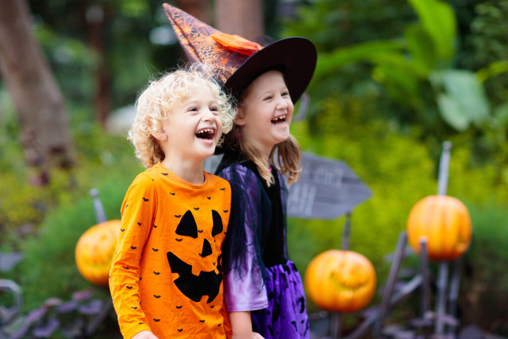 A brother and sister enjoy halloween. He is wearing a carved pumpkin themed t-shirt and she is dressed as a witch. They are laughing at something out of frame.