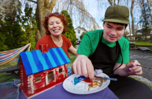 A teenage son and his mother sitting outside painting a small wooden house on a tabletop. They are both smiling, while the son dips his fingers into the paint tray.