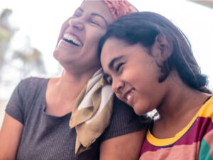 A young teenage girl leans her head on her Mum's shoulder. Her eyes are closed, and she smiles. Her mum wears a bandana and laughs.