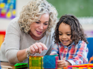 A kindergarten teacher plays and interacts with a happy indigenous boy. They sit at a table, build with colourful shapes and enjoy each other's company.