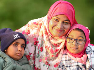 A mother in a hajib is sitting in the garden with her young son on the left and daughter on the right. Her son looks into the distance and the Mum and daughter, who wears glasses, smile at the camera. She has her arms around both of her children.