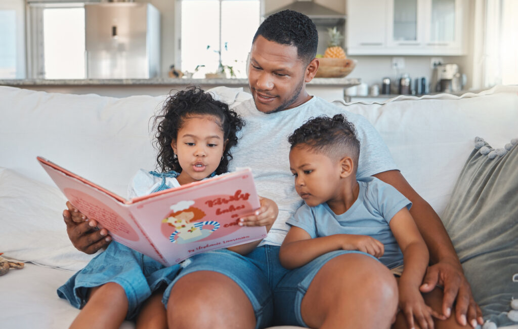 A young father, his daughter and son all read a picture book on the couch in their home.