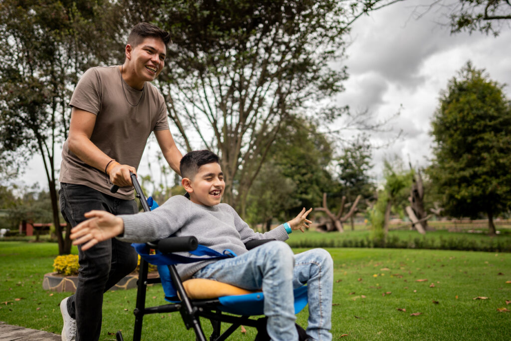 A boy in a wheelchair is being pushed by his dad through a park, he is smiling with his eyes closed and arms out wide. They are having fun.