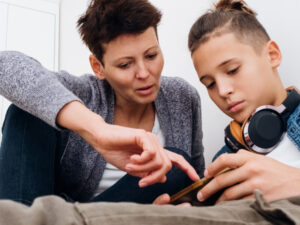 A teenage boy wearing headphones sits on his bed with his Mum. They both look at a mobile phone and his Mum points to the screen to explain what they are seeing.