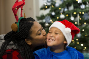 A mother kisses her smiling son on the cheek. She is wearing felt reindeer antlers and he is wearing a santa hat. They are in front of a christmas tree.