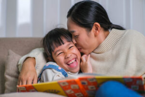 Mother reading a book with her daughter on the sofa at home. Happy mother reading a book, relaxing, hugging, telling funny stories to children and spending time together at home.