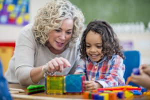 A kindergarten teacher plays and interacts with a happy, indigenous preschool boy. They sit at a table, build with colourful shapes and enjoy each other’s company.