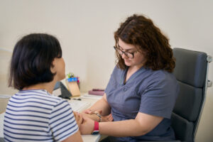 Alt text: A female doctor in a grey uniform sits at a desk, gently holding the hands of a female patient in a striped shirt. They are in a medical office.